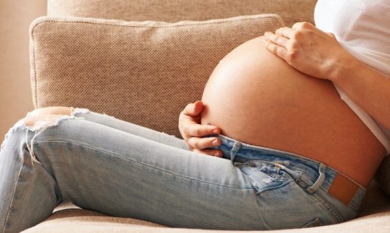 A pregnant woman sitting down on a tan couch with her hands around her stomach.