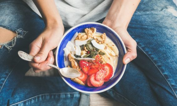 Yogurt, granola, seeds, fresh and dry fruits and honey in blue ceramic bowl in woman' s hands.