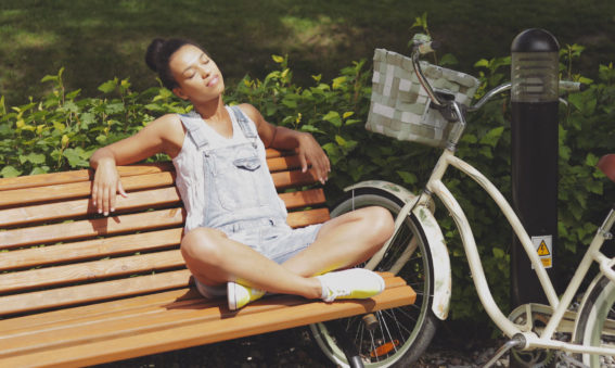 Young casual woman sitting with legs crossed and eyes closed on wooden bench in park with bicycle near and enjoying sunlight.